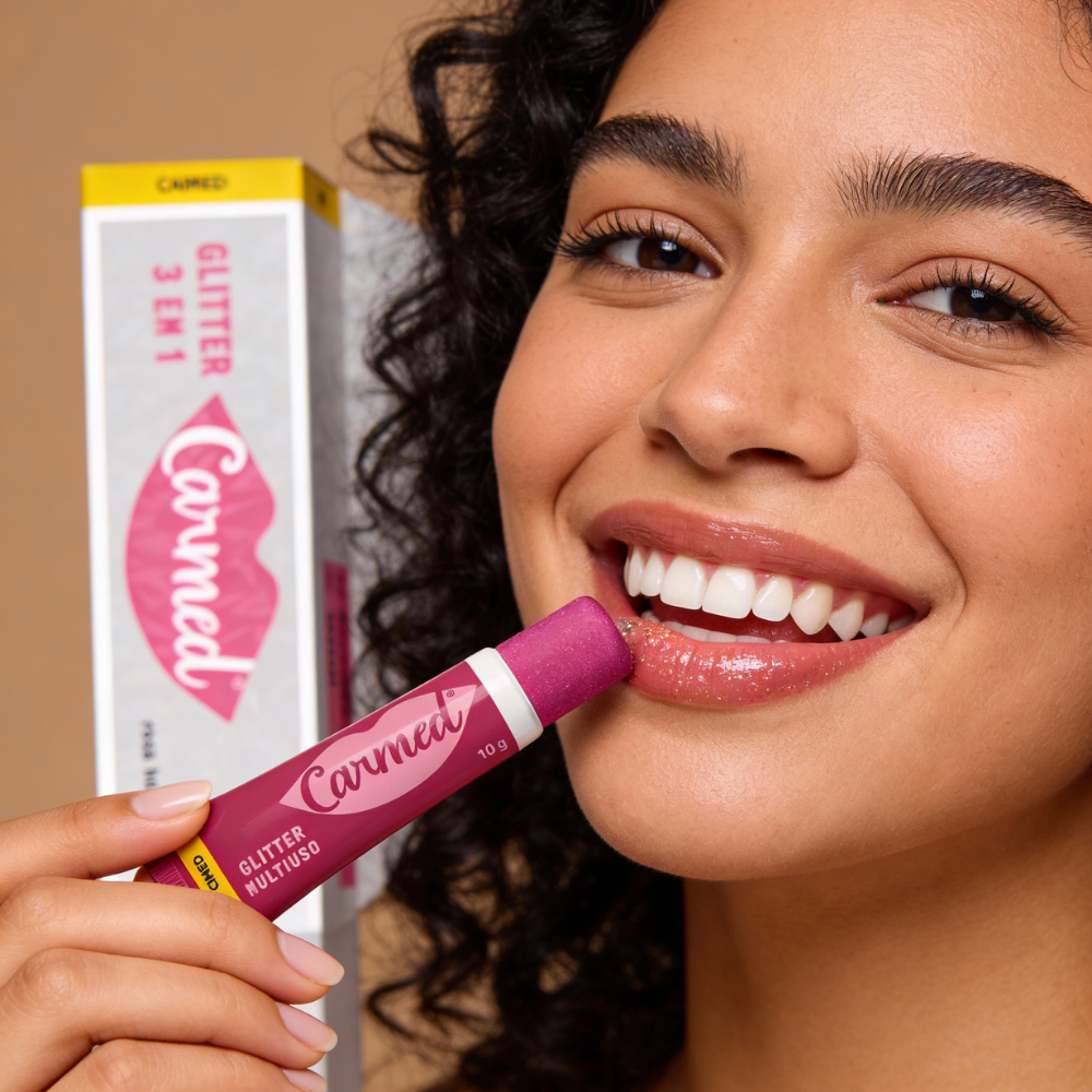 A smiling woman holds a tube of Carmed Glitter Multiuso 3 em 1 lip balm, showcasing its vibrant packaging.