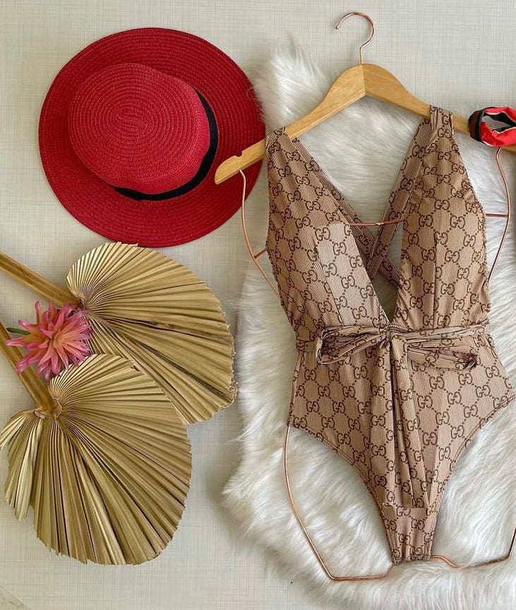 A beige swimsuit with a Gucci-inspired pattern displayed on a mannequin, accompanied by a red hat and a decorative palm fan.