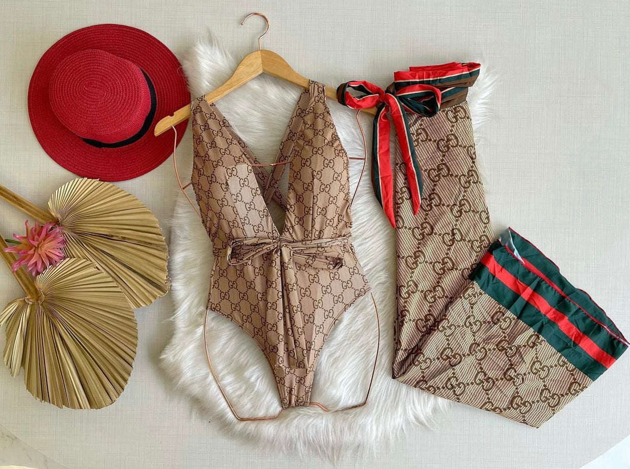A beige swimsuit with a Gucci-inspired pattern displayed on a mannequin, accompanied by a red hat and a decorative palm fan.