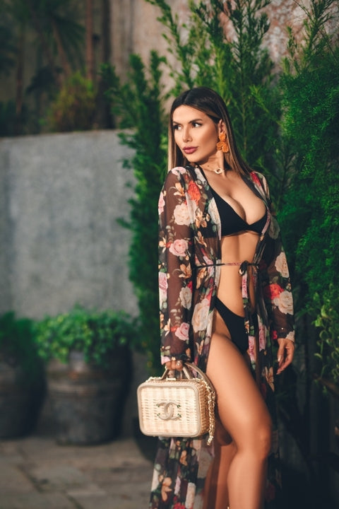 This image shows a woman posing in a black bikini with a sheer floral cover-up. She is also carrying a quilted handbag featuring the iconic double C logo, which is characteristic of the luxury fashion house Chanel.