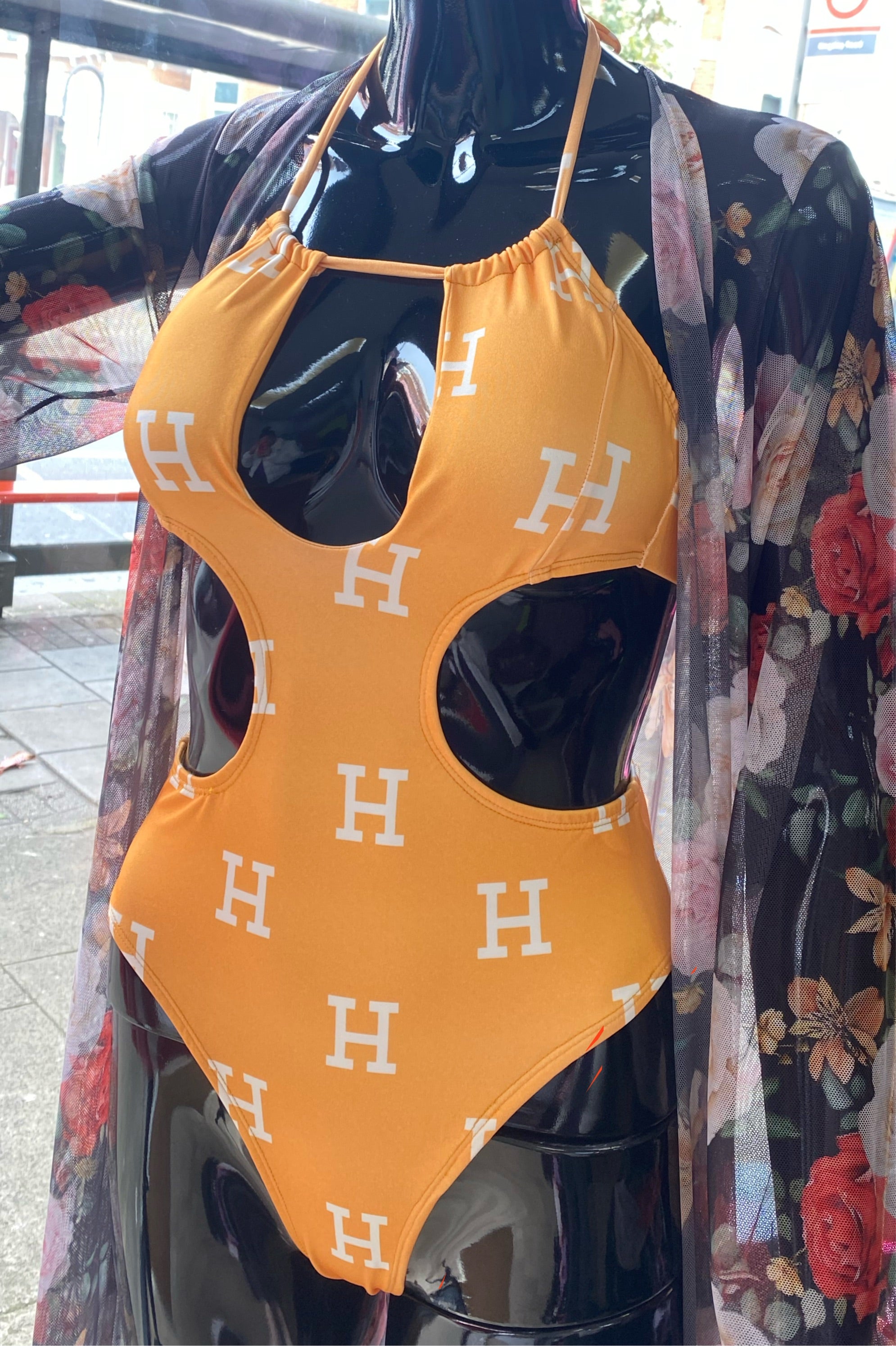 An orange one-piece swimsuit with a floral pattern and the letter 'H' repeated throughout, displayed on a mannequin.