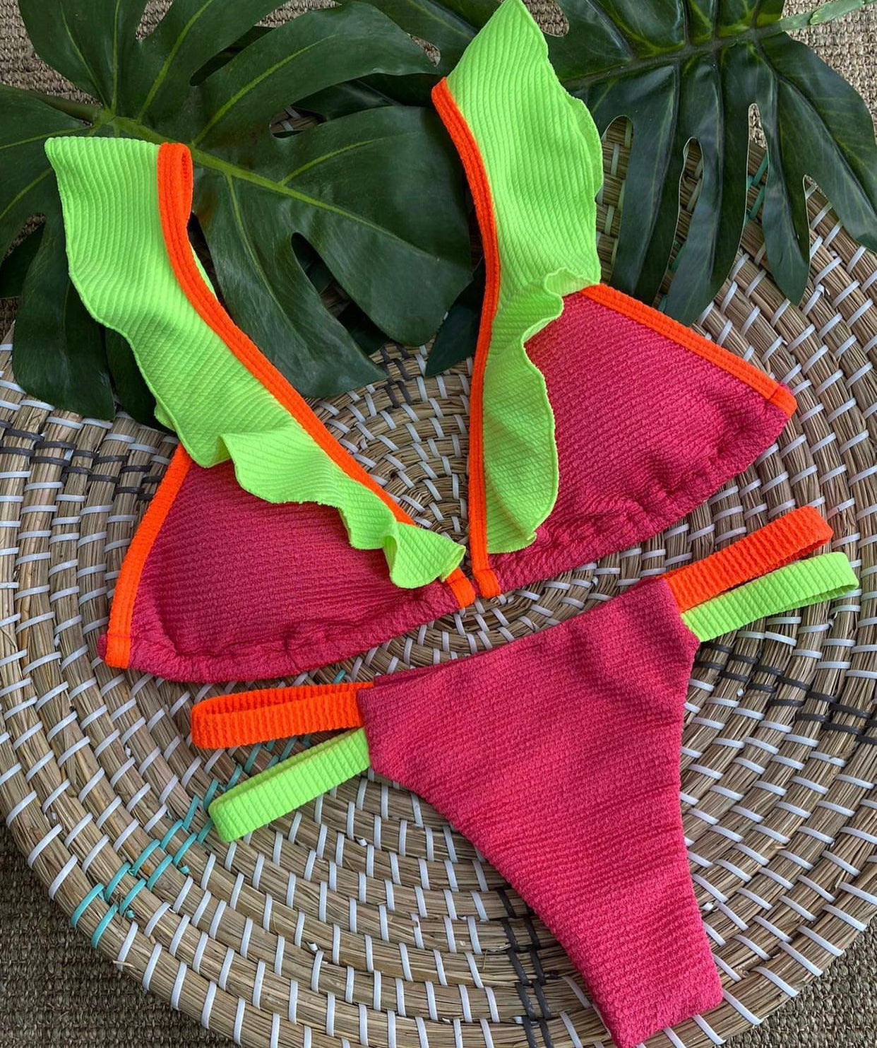 A bikini with pink and green fringe details, displayed on a woven mat.