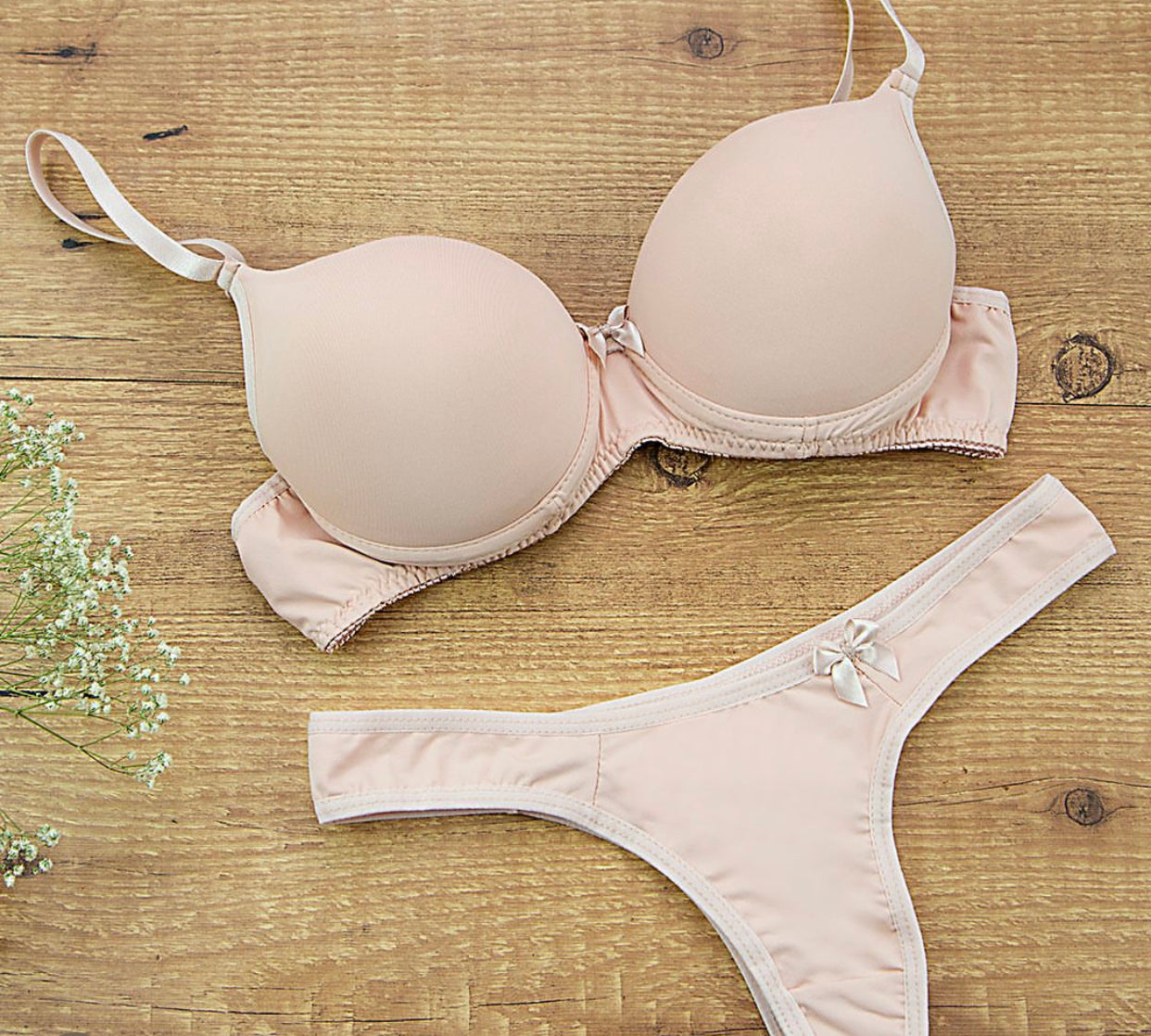 A plain beige bra and panty set on a wooden surface with a small flower arrangement to the side.